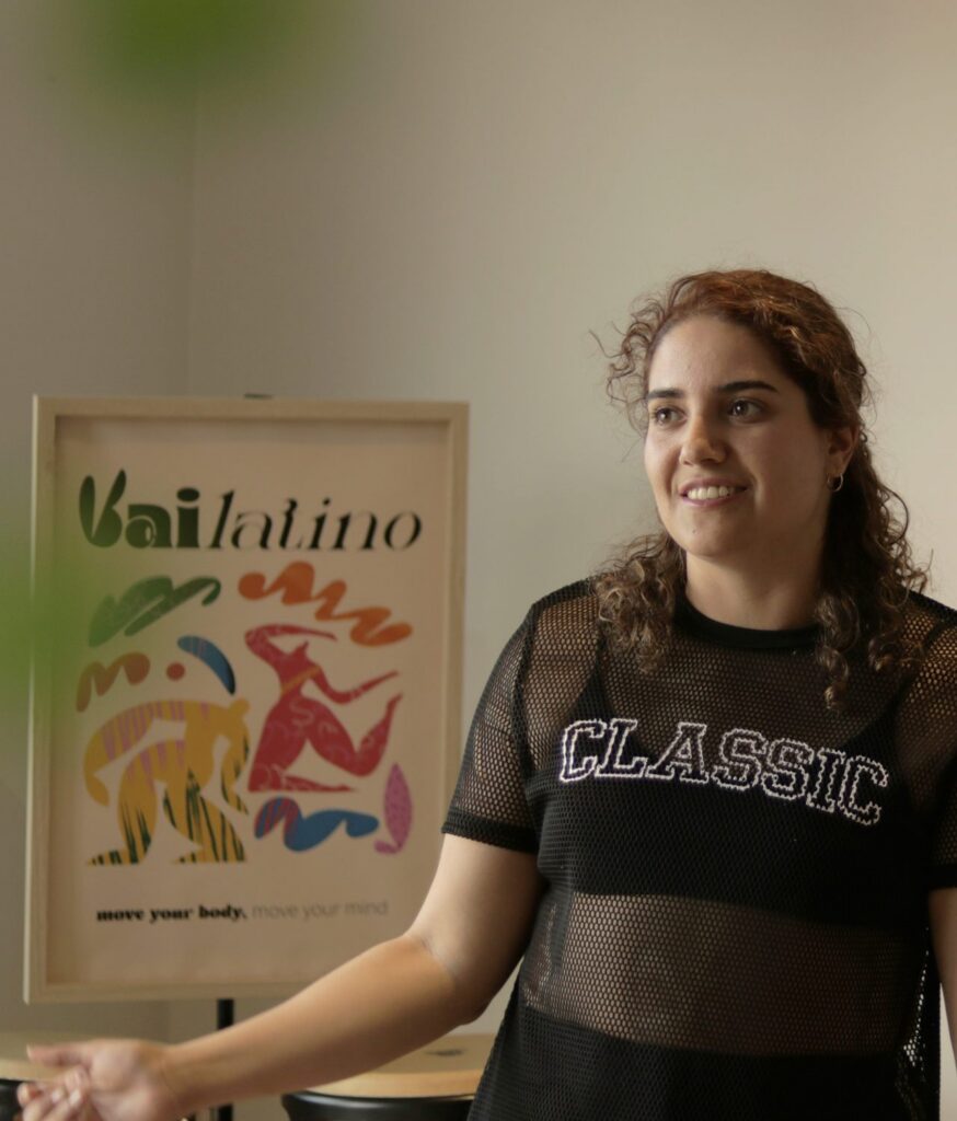 Vanessa smiles as she explains a step, beside the Vailatino poster